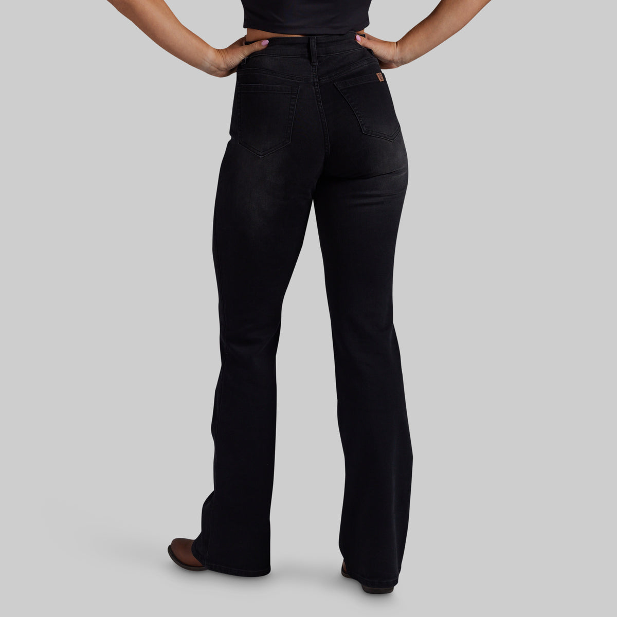 Rear view of a person wearing black mid-rise bootcut jeans against a light gray studio background.