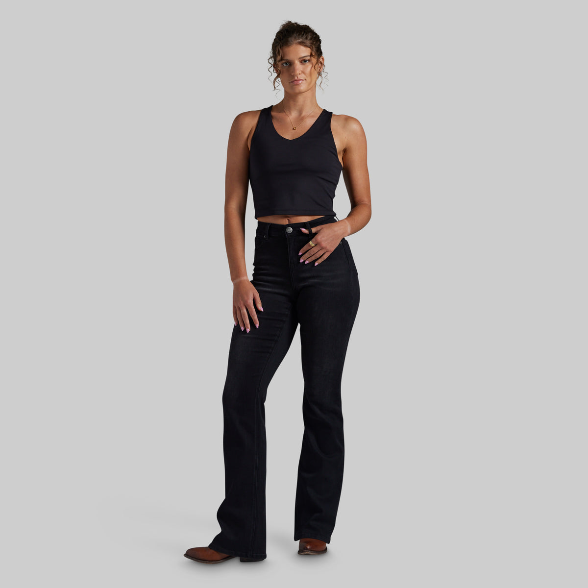 A woman modeling a black sleeveless crop top and black mid-waisted bootcut jeans against a solid light gray background.