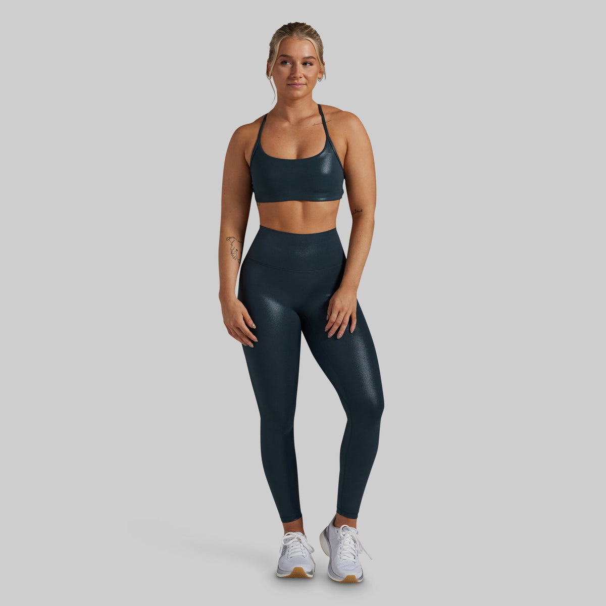 A woman modeling a matching dark teal athletic set consisting of a sports bra and high-waisted leggings, paired with white sneakers, against a solid light gray background.