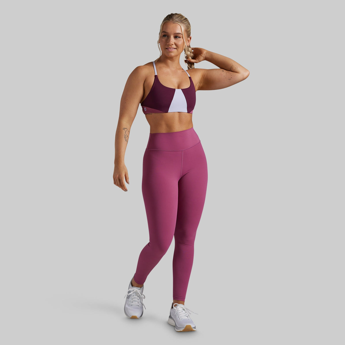 A woman modeling a color-blocked sports bra and matching high-waisted leggings in a studio setting with a solid gray background.