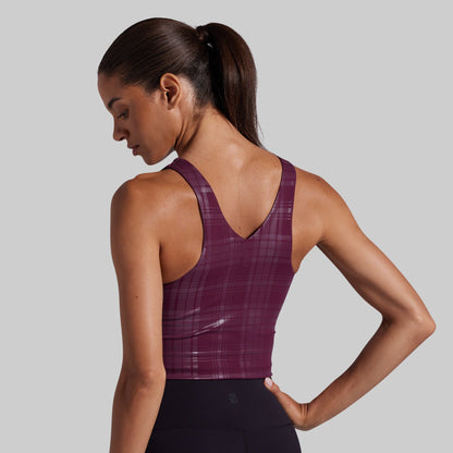 A woman shown from the back wearing a plum purple athletic sports bra with a subtle plaid pattern, paired with black leggings, against a solid gray studio background.