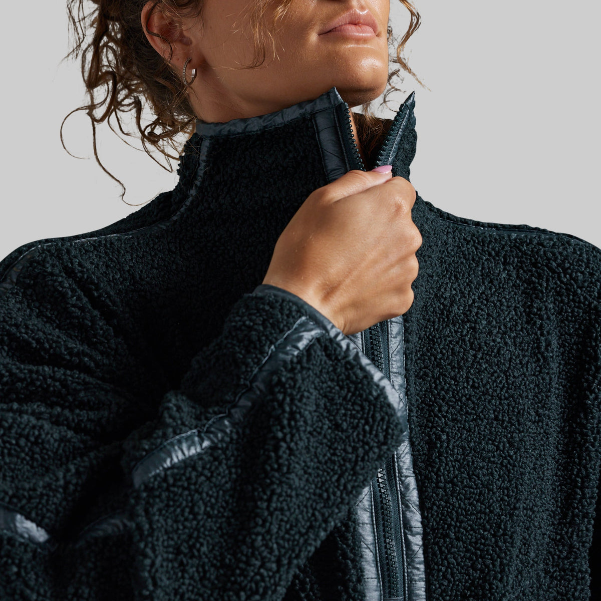 Close-up of a person wearing a dark teal fleece zip-up jacket, pulling the zipper with one hand against a plain light gray background.