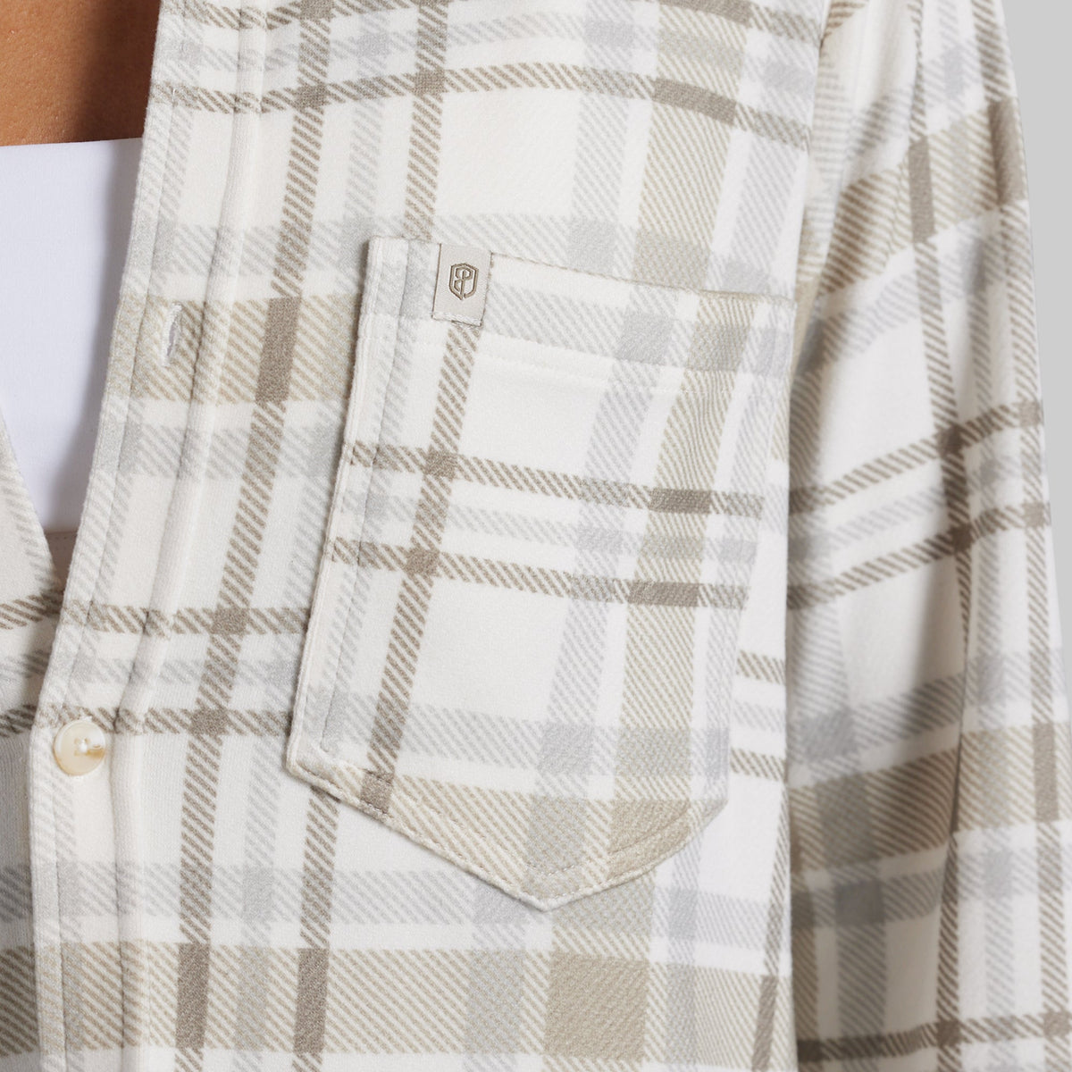 Close-up of a plaid button-up shirt with a chest pocket, featuring a cream and beige pattern with a small logo patch on the pocket.