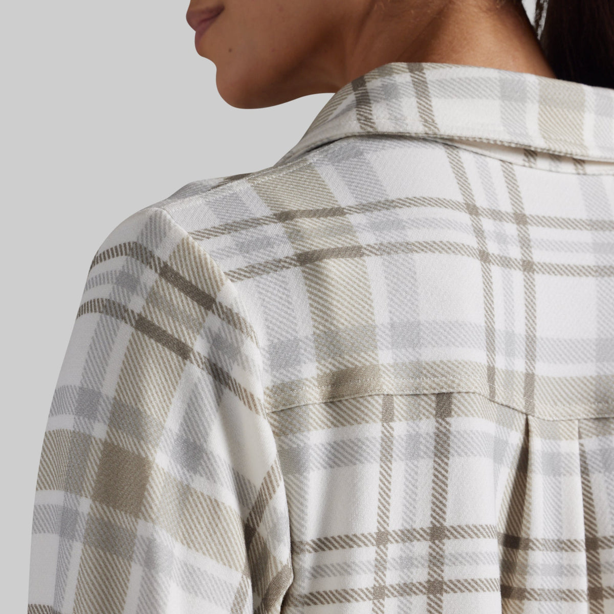 Close-up of a person wearing a plaid button-up shirt, showing the back of the neck, shoulder, and upper back against a light gray studio background.