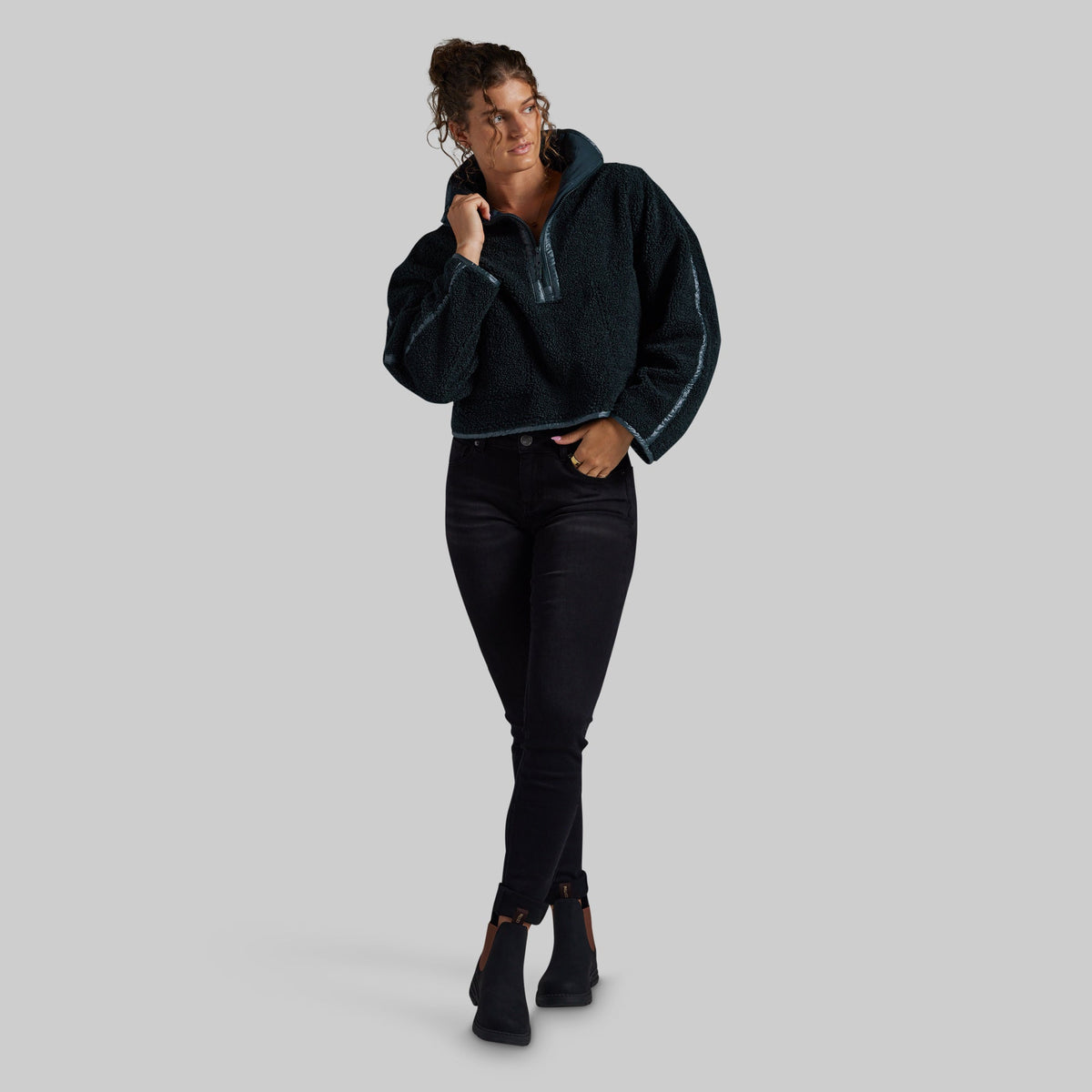 A woman modeling a dark teal fuzzy quarter-zip hoodie with contrasting light piping, paired with black skinny jeans and black ankle boots, against a solid gray background.