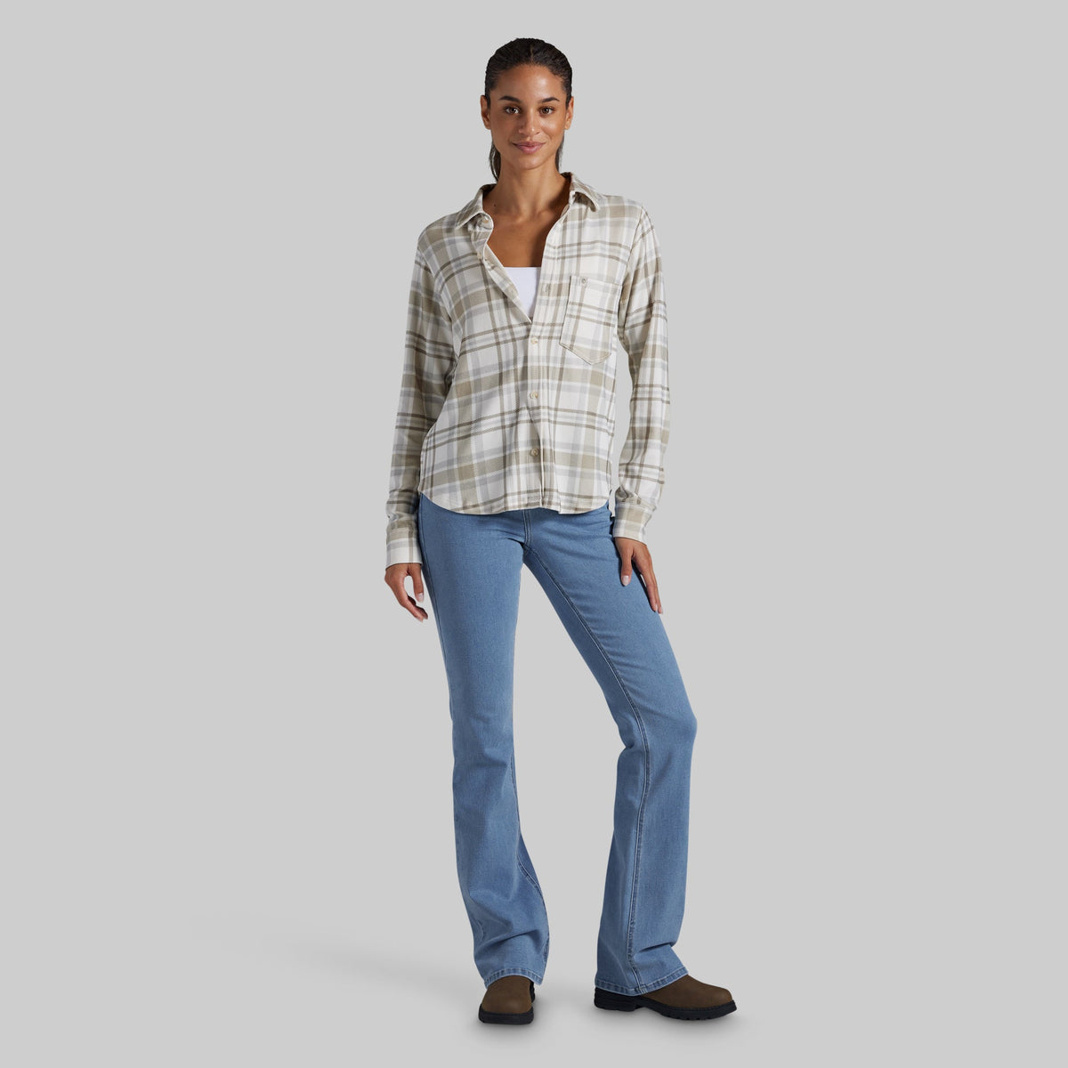 A woman modeling a long-sleeve plaid button-up shirt in beige and white with blue jeans and brown shoes, standing against a solid gray background.