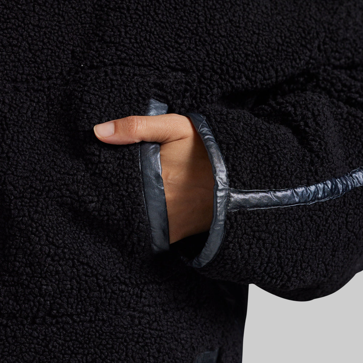 Close-up of a black sherpa fleece jacket with a hand tucked into a pocket, showing a contrasting dark gray shiny trim along the pocket opening.