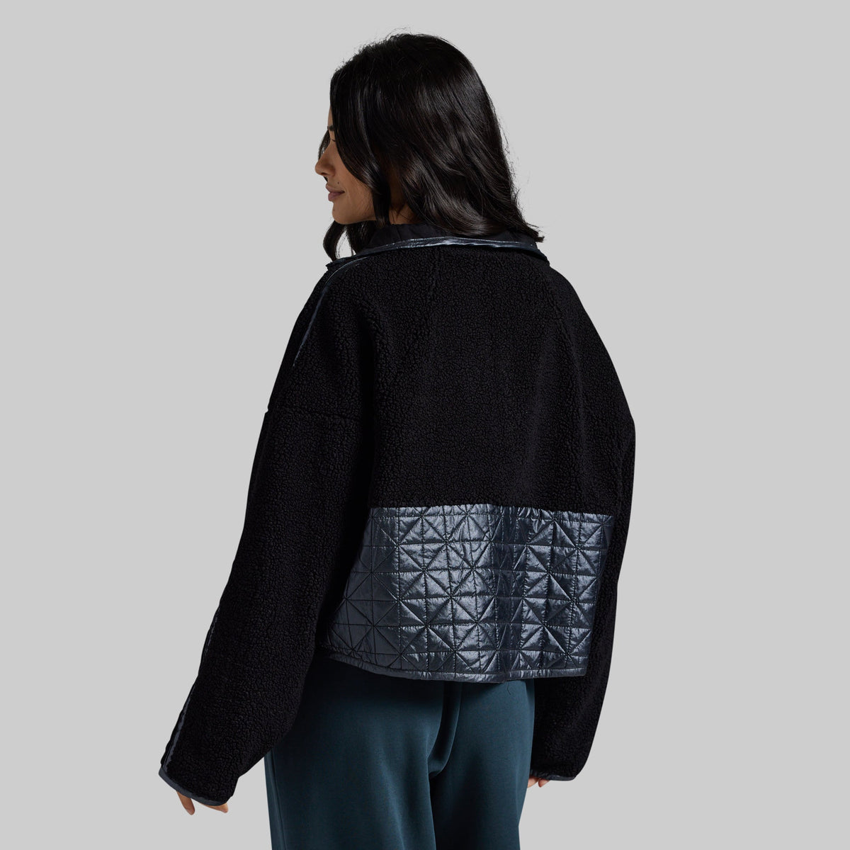 A woman shown from the back wearing a black cropped jacket with a dark gray quilted geometric patterned panel on the lower back, paired with dark teal pants, against a solid light gray background.