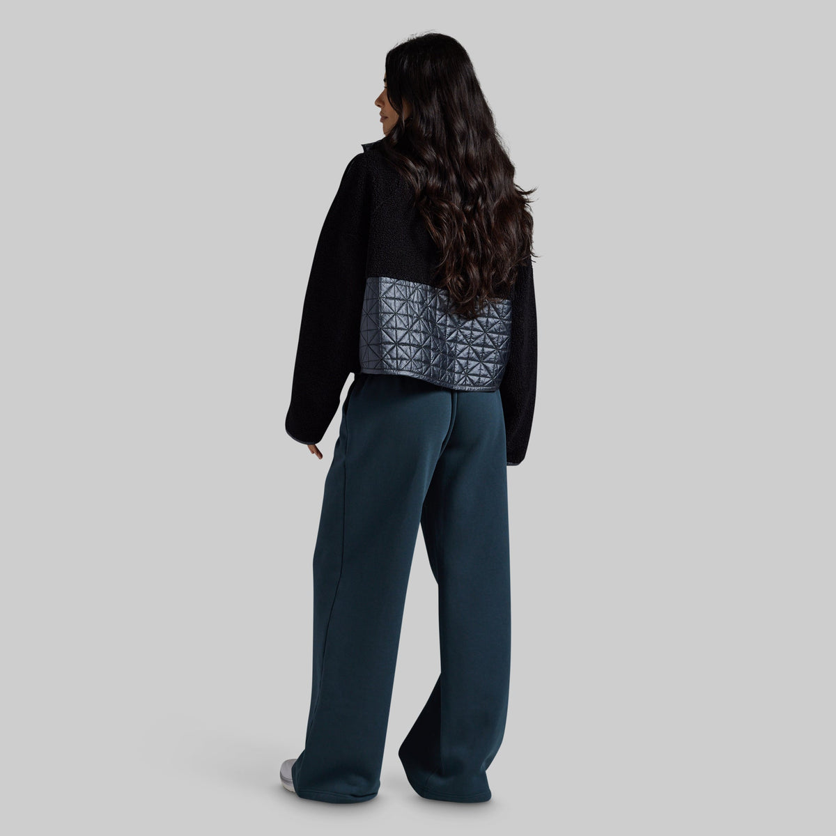 A woman is shown from behind wearing a black cropped jacket with a textured gray geometric quilted waist panel, paired with dark teal wide-leg trousers, against a solid gray background.