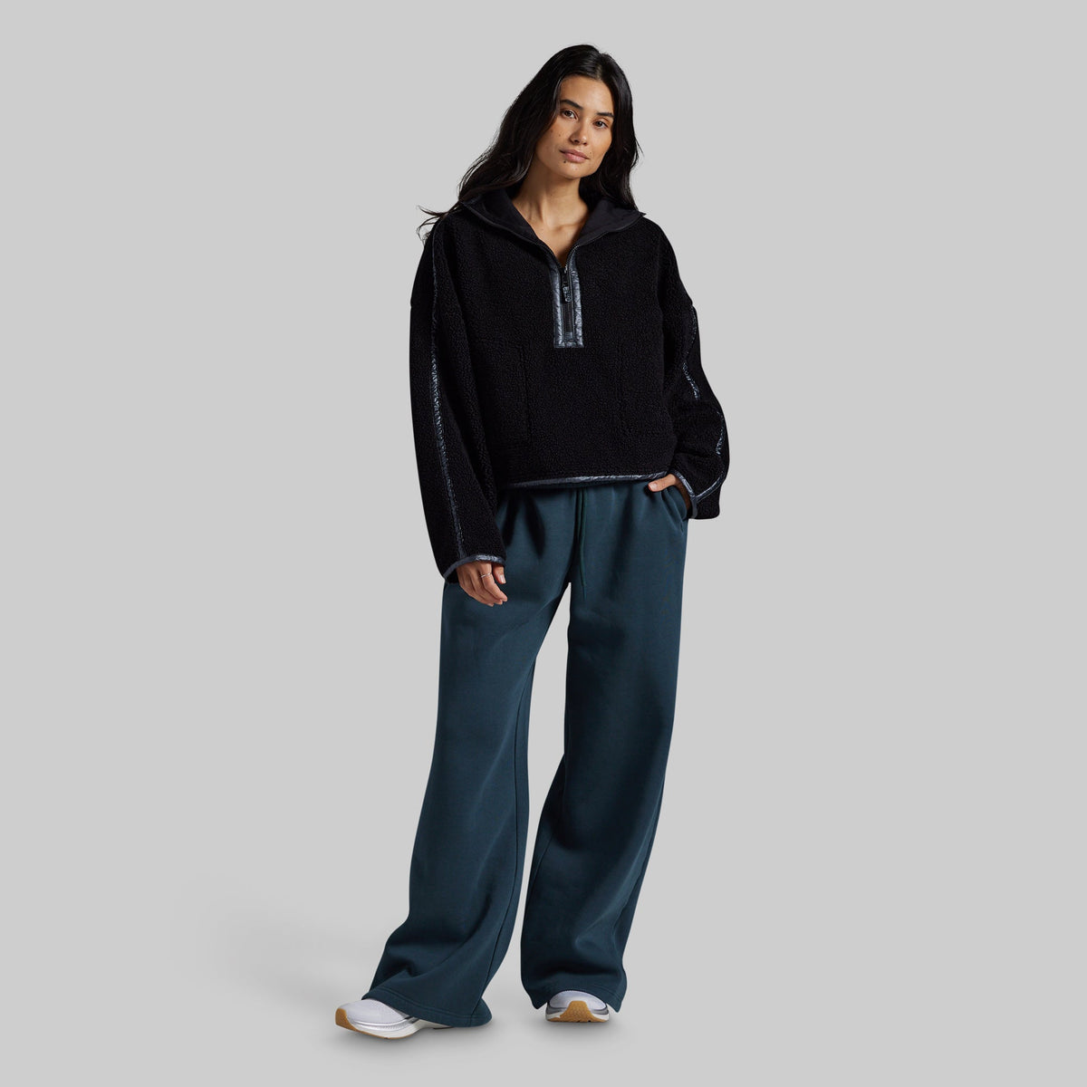 A woman modeling a black fleece half-zip pullover with textured knit sleeves and dark teal wide-leg sweatpants, standing against a solid gray studio background.