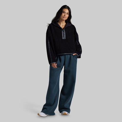 A woman modeling a black fleece half-zip pullover with textured knit sleeves and dark teal wide-leg sweatpants, standing against a solid gray studio background.
