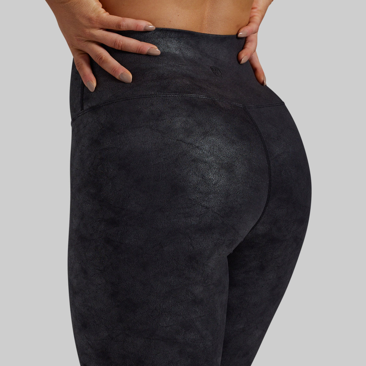Close-up back view of a person wearing dark charcoal high-waisted leggings against a light gray studio background.