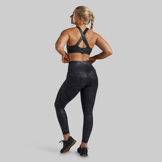 A woman shown from the back wearing a matching black athletic set, featuring a sports bra with crisscross straps and high-waisted leggings with a subtle mottled pattern, set against a solid gray background.