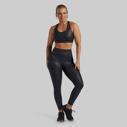 A woman modeling a matching dark gray and black textured activewear set, consisting of a sports bra and high-waisted leggings, against a solid gray background.