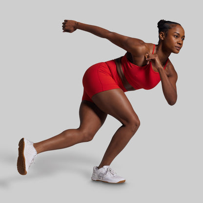 Woman in red athletic outfit and white sneakers posing in a running stance on a light gray background