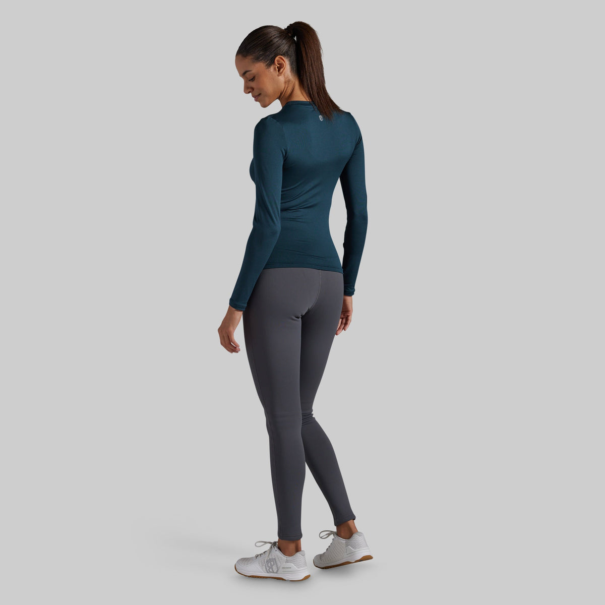 A woman wearing a dark teal long-sleeve athletic top and dark gray leggings, shown from the back against a solid light gray background.