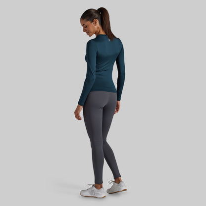 A woman wearing a dark teal long-sleeve athletic top and dark gray leggings, shown from the back against a solid light gray background.
