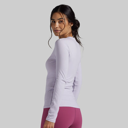 A woman wearing a fitted long-sleeve athletic top in a very light lavender-purple color, paired with magenta leggings, shown from a side-back angle against a solid light gray studio background.