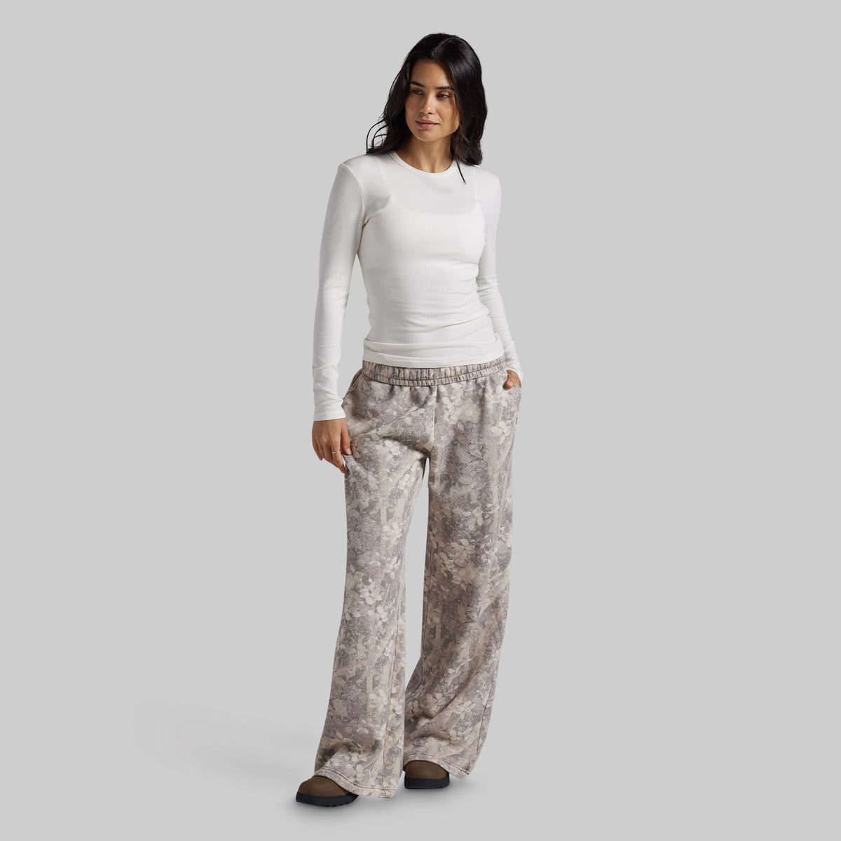 A woman modeling a white long-sleeve crewneck top and loose, wide-leg sweatpants with a beige and gray abstract floral pattern, set against a solid light gray background.