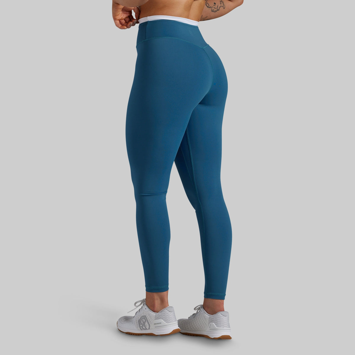 A person wearing high-waisted teal blue leggings, shown from the back and side against a light gray studio background. The leggings are fitted and extend to the ankles, with a white waistband visible above the hips.