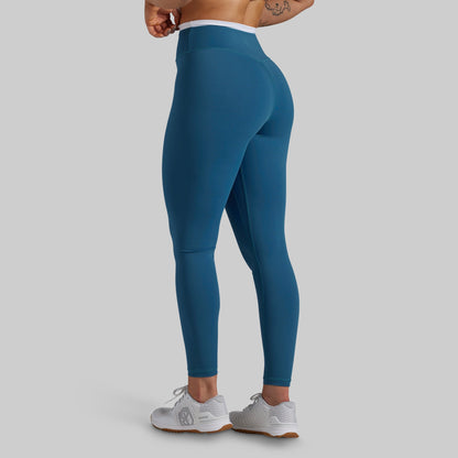A person wearing high-waisted teal blue leggings, shown from the back and side against a light gray studio background. The leggings are fitted and extend to the ankles, with a white waistband visible above the hips.