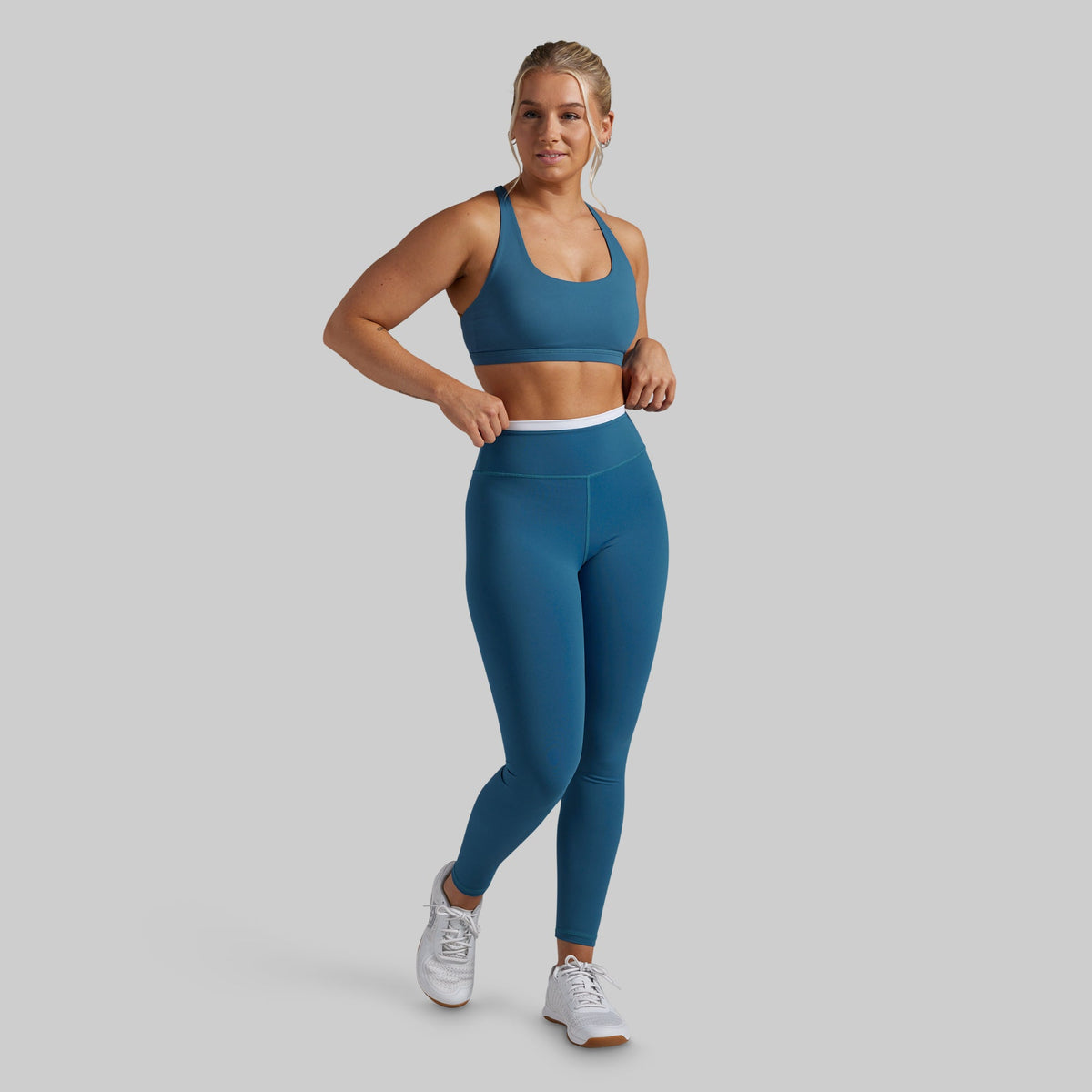 A woman modeling a matching athletic set consisting of a teal blue sports bra and high-waisted leggings with a white waistband, paired with white sneakers, against a solid gray background.