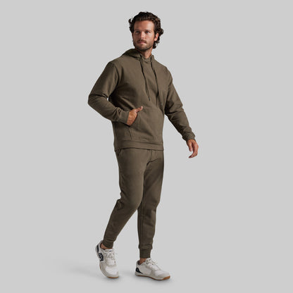 A man modeling a matching olive green hoodie and jogger sweatpants set, paired with white sneakers, against a solid light gray background.