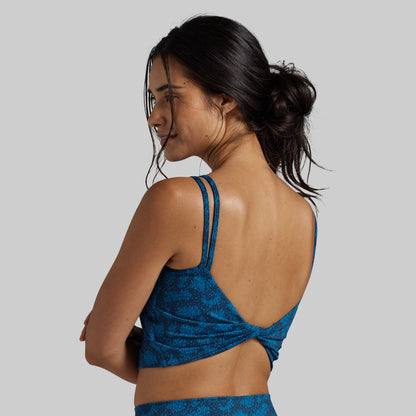 A woman shown from the back wearing a blue patterned sports bra with double shoulder straps and a twisted back detail, against a solid gray background.