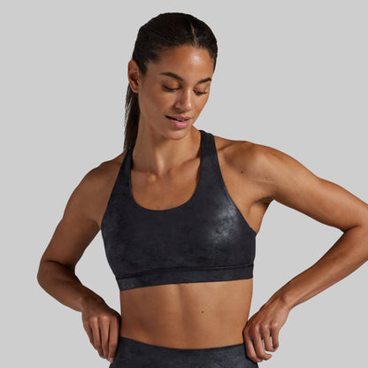 A woman wearing a marbled black athletic sports bra with a subtle textured pattern, shown from the chest up against a solid light gray background.