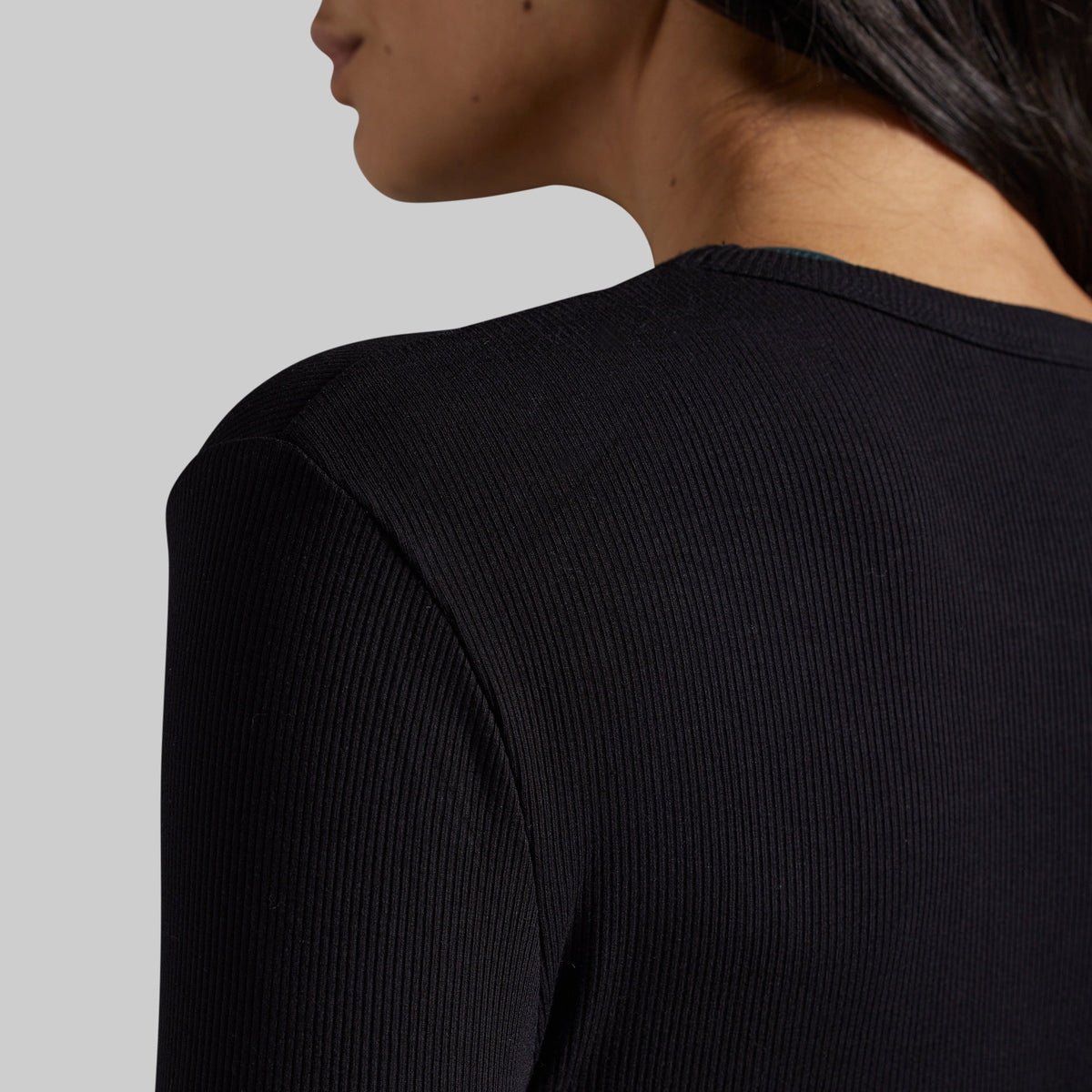 Close-up of a person wearing a black ribbed long-sleeve top, showing the back and shoulder against a light gray studio background.