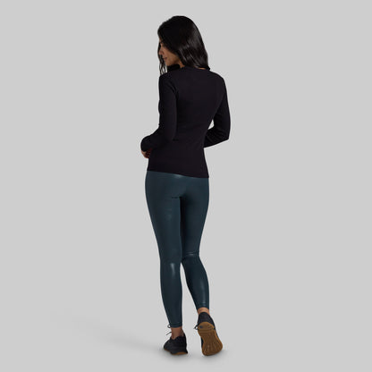 A woman is shown from the back wearing a black long-sleeve top and dark teal shiny leggings, standing against a solid light gray background.
