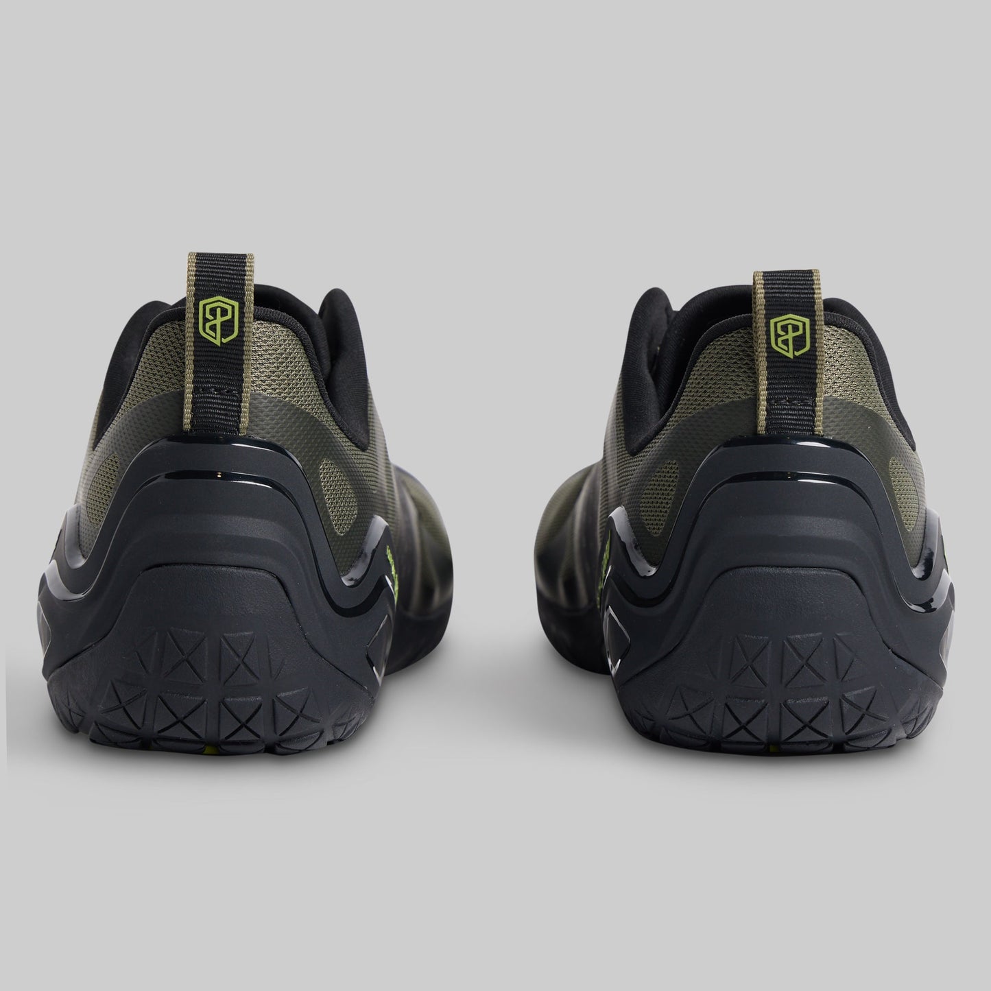 A pair of athletic sneakers shown from the rear, featuring a black and olive green design with a small yellow-green shield logo on the heel pull tabs, set against a solid gray background.