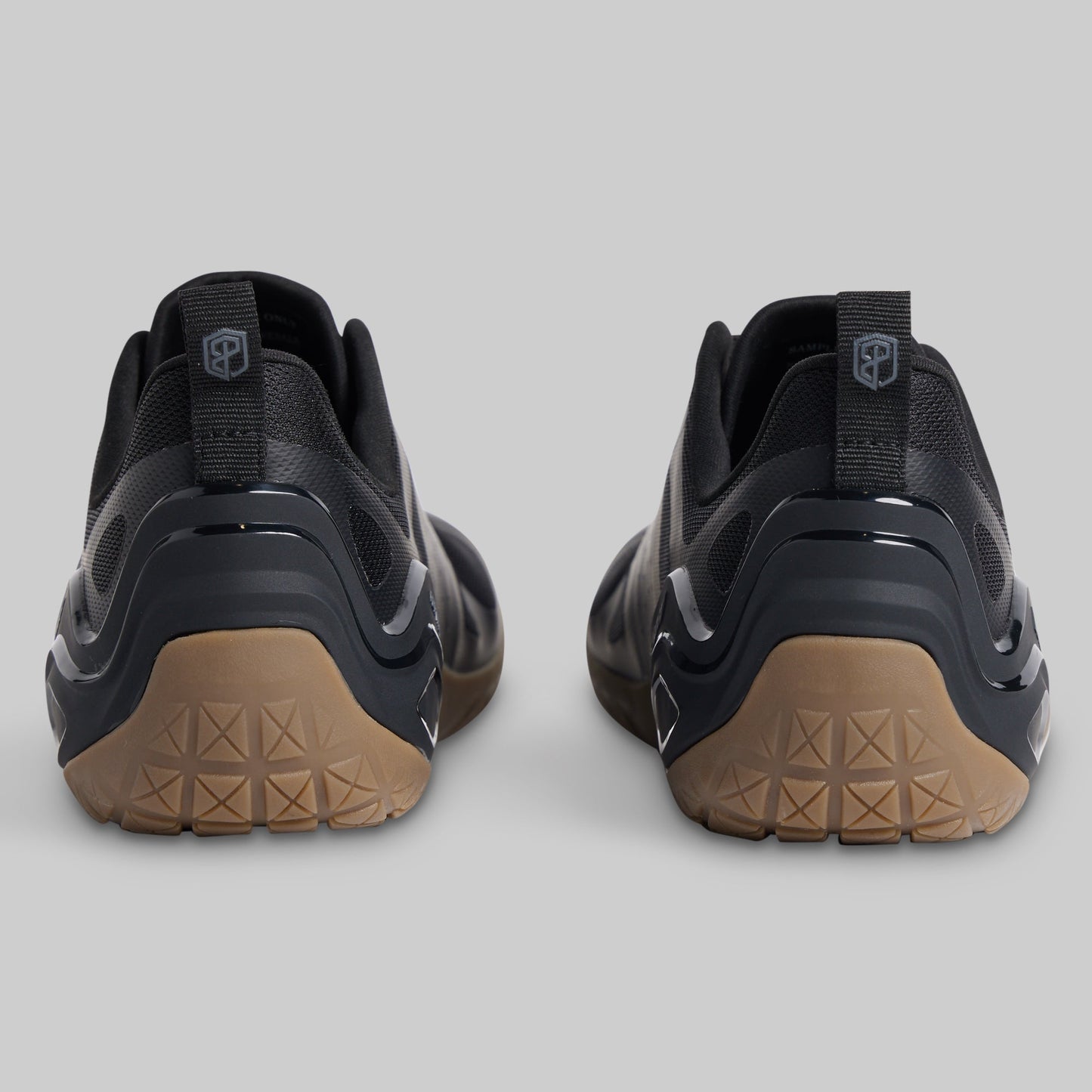 A pair of black minimalist sneakers shown from the rear, featuring textured tan outsoles and a pull tab with a shield logo on a light gray background.