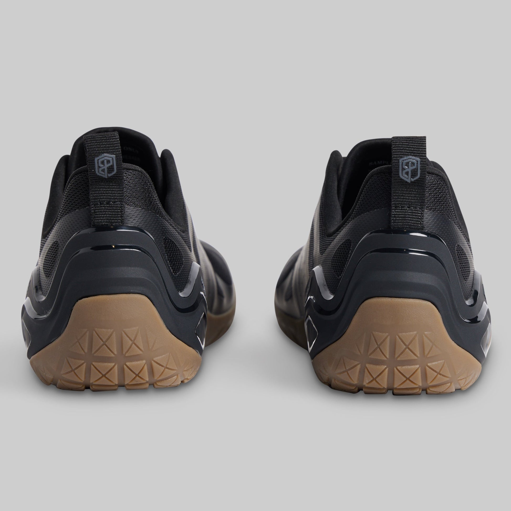 A pair of black minimalist sneakers shown from the rear, featuring textured tan outsoles and a pull tab with a shield logo on a light gray background.