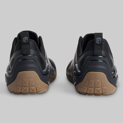 A pair of black minimalist sneakers shown from the rear, featuring textured tan outsoles and a pull tab with a shield logo on a light gray background.