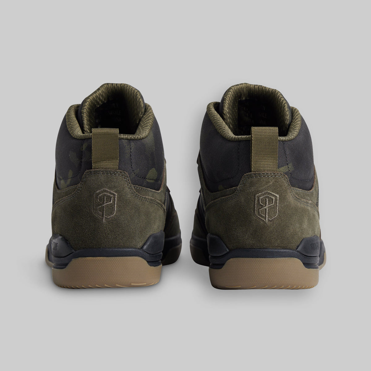 A pair of mid-top sneakers shown from the back, featuring olive green suede and camo panels, dark navy overlays, and tan rubber soles, set against a solid gray background.