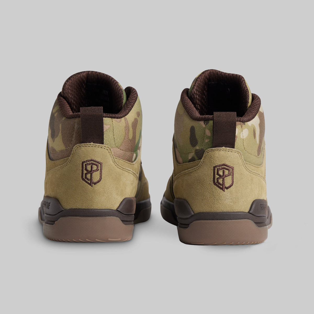 A pair of mid-top sneakers shown from the back, featuring a tan and brown camouflage pattern, suede-like tan panels, dark brown pull tabs, and a dark brown sole, set against a solid gray background.