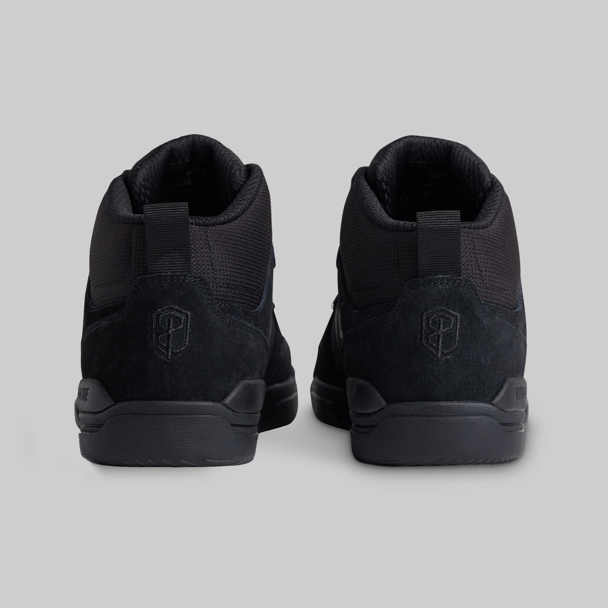 A pair of black mid-top sneakers shown from the back, featuring a textured mesh and suede upper with a pull tab and a shield logo on the heel, set against a solid gray background.
