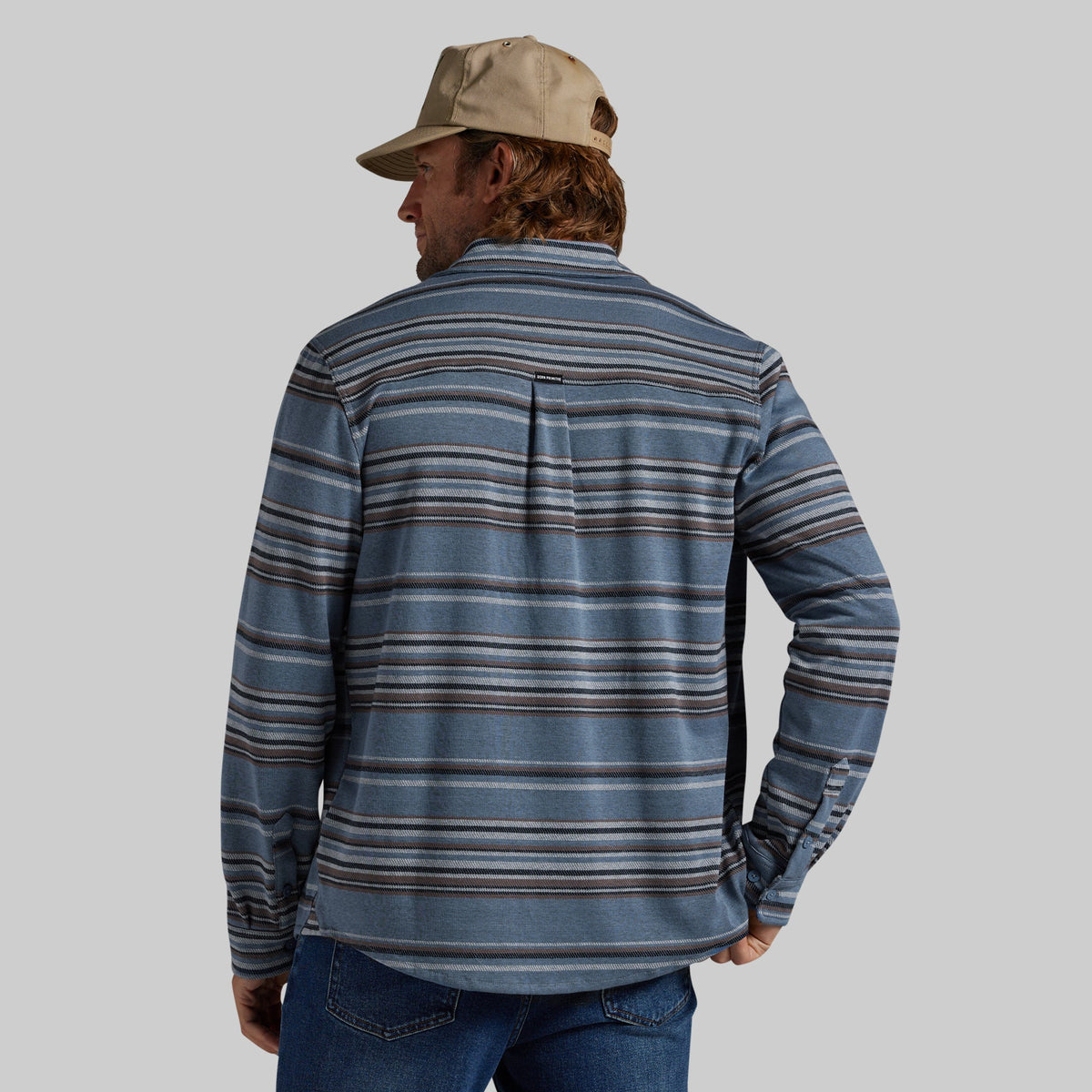 A man wearing a blue striped long-sleeve shirt and a tan baseball cap, photographed from the back against a solid gray background.