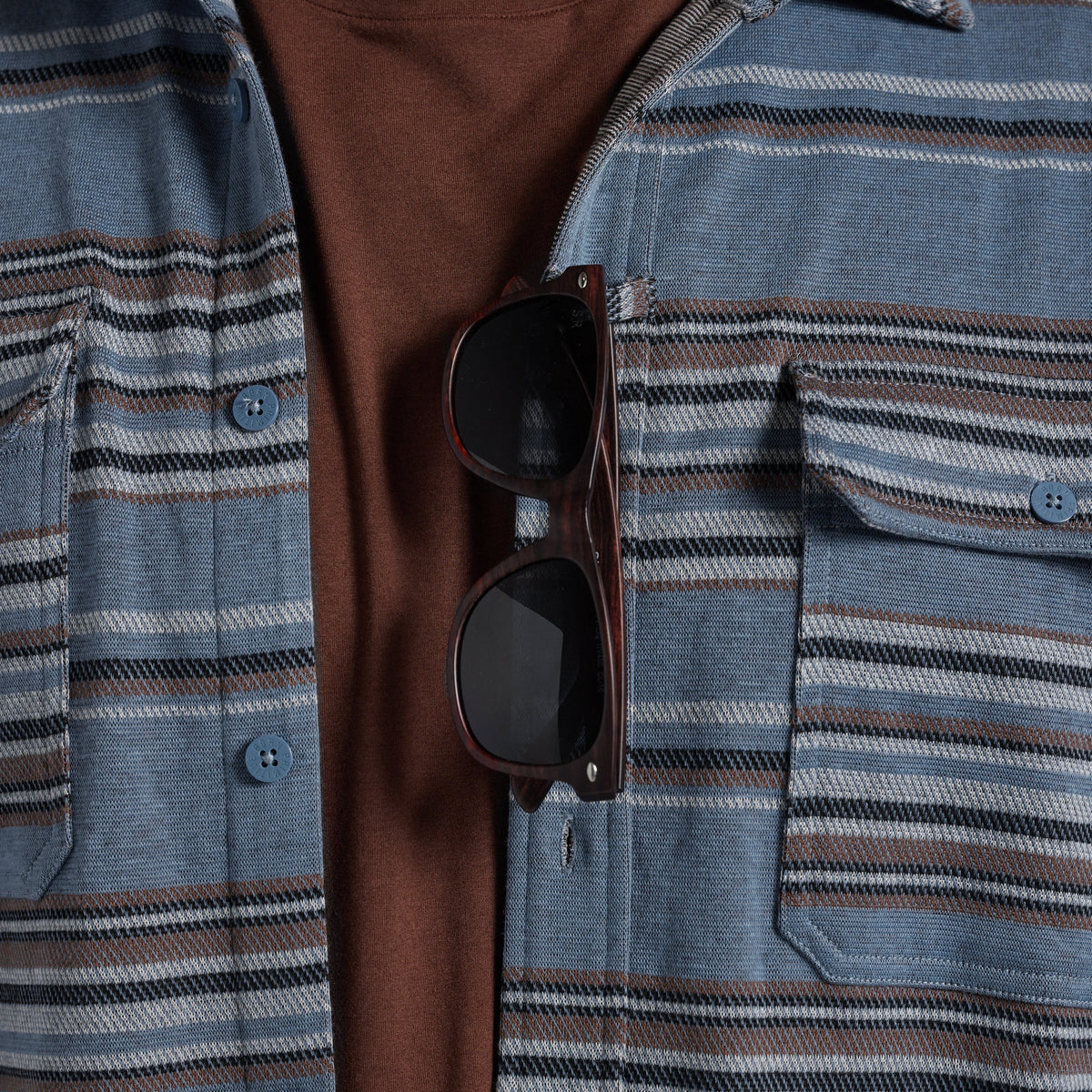 Close-up of a person wearing a blue striped button-up shirt over a brown t-shirt, with dark sunglasses hanging from the sunglass loop.
