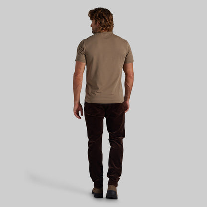 A man shown from behind wearing a short-sleeve taupe t-shirt and dark brown corduroy trousers, standing against a solid light gray background.