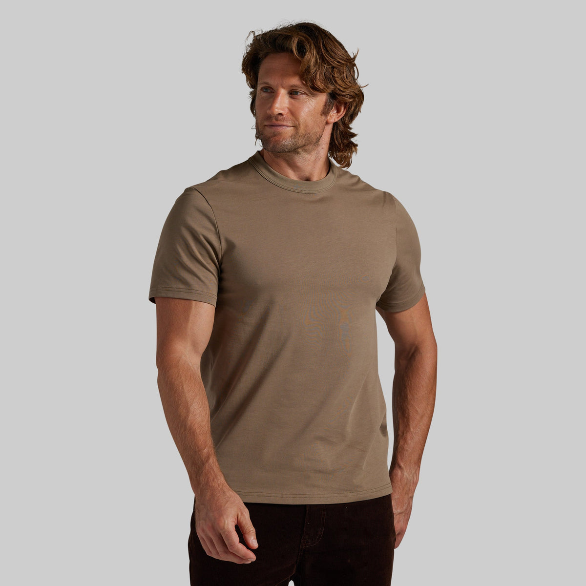 A man wearing a short-sleeve crewneck t-shirt in a muted taupe-brown color, paired with dark pants, standing against a solid light gray background.