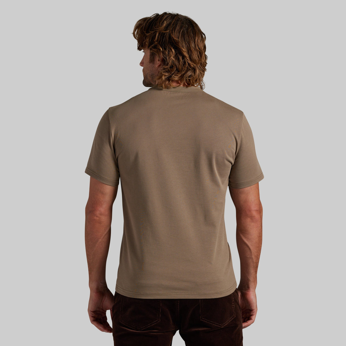 A man viewed from behind wearing a plain taupe-brown short-sleeve t-shirt and dark brown corduroy pants, set against a solid light gray background.