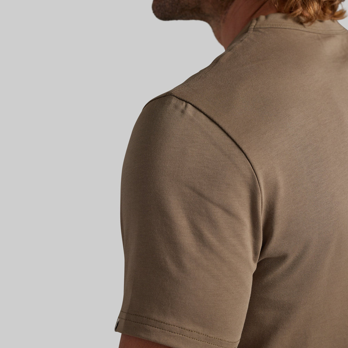 Close-up of a man wearing a short-sleeve crew-neck t-shirt in a warm taupe-brown color, showing the back and shoulder detail against a solid light gray background.