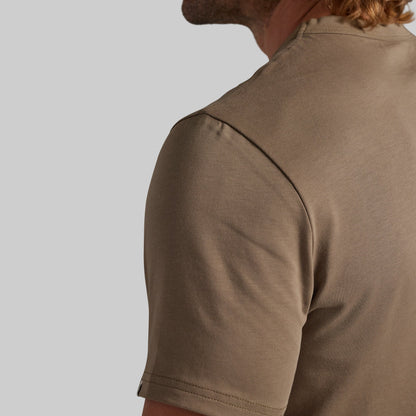 Close-up of a man wearing a short-sleeve crew-neck t-shirt in a warm taupe-brown color, showing the back and shoulder detail against a solid light gray background.