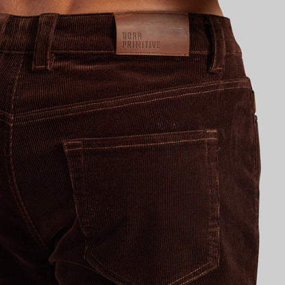 Close-up of the back waistband and pocket of dark brown corduroy pants, showing a brown leather patch with the brand name 'BORN PRIMITIVE'.