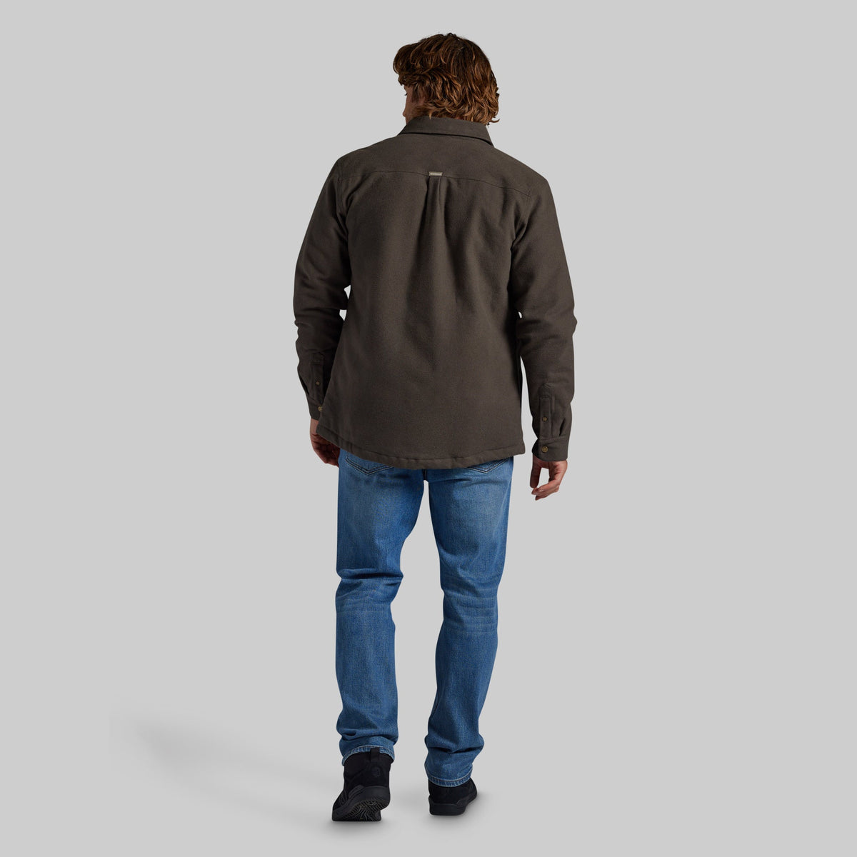 A man viewed from behind wearing a dark brown long-sleeve button-up shirt, blue jeans, and black shoes, standing against a solid gray studio background.
