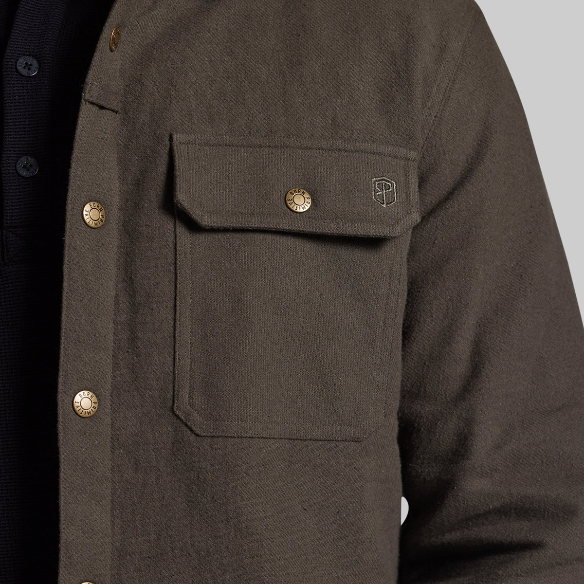 Close-up of a dark olive brown textured overshirt jacket with brass buttons and a small embroidered shield logo on the chest pocket, worn over a dark shirt.