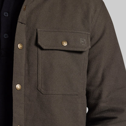 Close-up of a dark olive brown textured overshirt jacket with brass buttons and a small embroidered shield logo on the chest pocket, worn over a dark shirt.