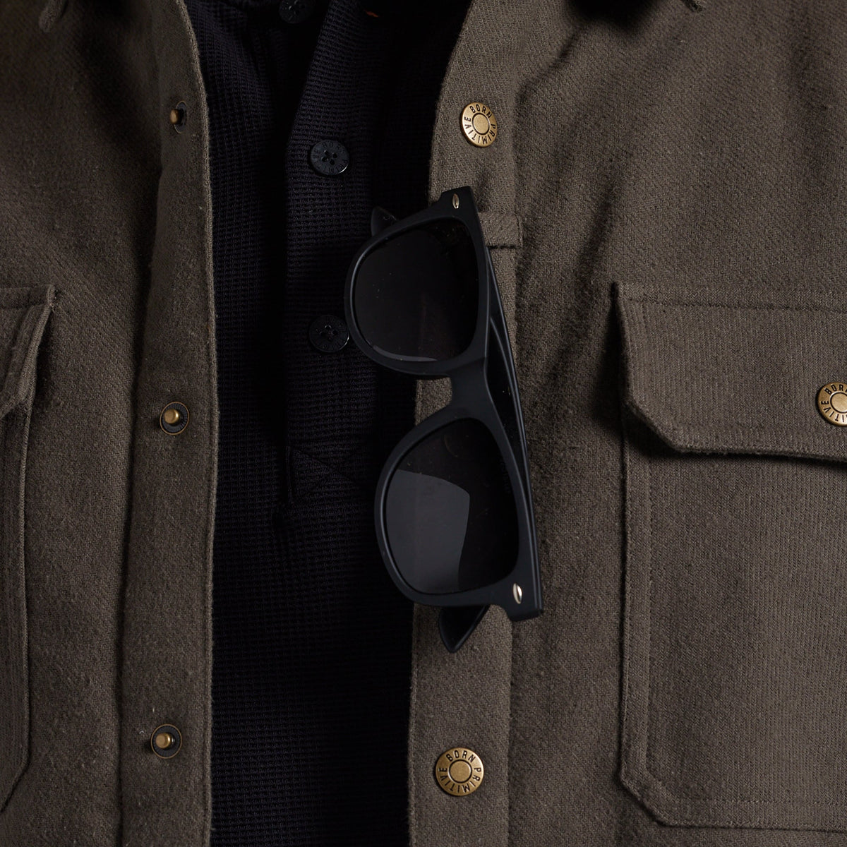 Close-up of a person wearing an olive brown textured jacket over a black knit shirt, with black sunglasses resting on the chest pocket.
